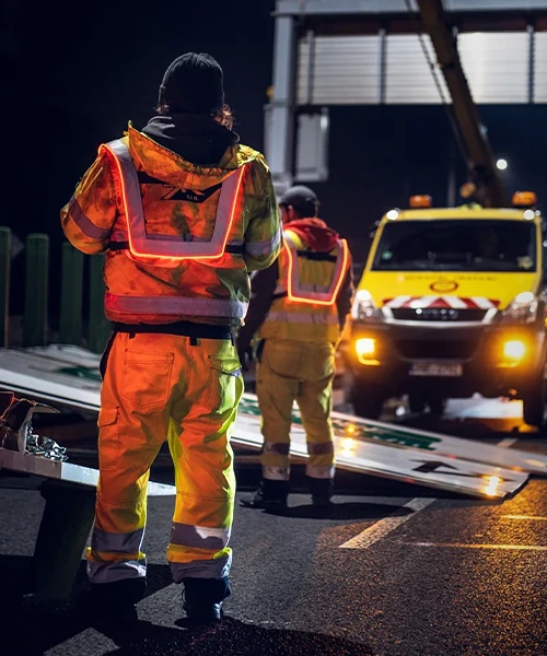 Trabajadores nocturnos reparando carreteras, vistos de espaldas con chalecos EquiRay Heavy Duty, cuyas fibras ópticas Sunfibre activadas brillan en la oscuridad mientras recogen señales dañadas