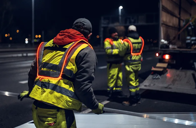 Trabajador reparando señales en la carretera de noche, visto de espaldas con su chaleco Heavy Duty, mientras más adelante, dos compañeros equipados también con chalecos conversan junto al camión